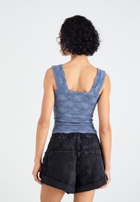 Modra pletena majica brez rokavov z teksturiranim cvetličnim vzorcem in širokimi naramnicami, v kombinaciji z visokim pasom črnimi denim kratkimi hlačami, s vidnimi podrobnostmi na hrbtu.