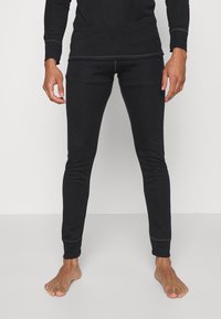 Leggings termici neri con un design aderente, realizzati in tessuto elastico. Rifiniti con cuciture piatte sui bordi per il comfort. Indossati a piedi nudi.