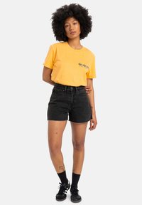 Yellow cotton t-shirt with a black graphic design, paired with black denim shorts. Model wearing black socks and shoes, standing against a white background.