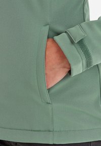 Green jacket detail showing a side pocket and cuff with a soft texture. Features include stitching accents for durability and functionality.