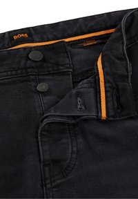 Černé tapered-fit džíny vyrobené z denimu, s dvěma černými knoflíky, oranžovými švy a jemnými známkami opotřebení na látce.