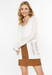 Cardigan bianco lavorato a maglia con apertura frontale, con un motivo in pizzo e maniche lunghe, abbinato a una gonna in suede marrone.