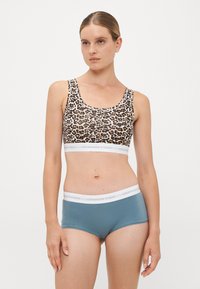 Kvinde med hår i en knold iført en leopardmønstret sports-bh og blå boyshorts undertøj med hvide elastiske bånd foran en ensfarvet baggrund.