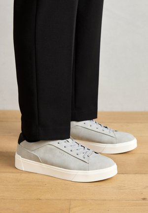Baskets en suède gris clair avec une finition texturée, semelles en caoutchouc blanc et accents de talon en cuir noir, associées à un pantalon noir ajusté.