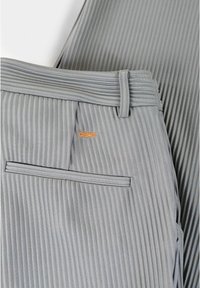 Graue, gefaltete Hose mit glatter Textur, einem rechteckigen Bund und einem kleinen orangenen Logo auf der hinteren Tasche.