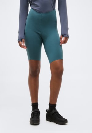 Άτομο που φοράει τιρκουάζ σορτς ποδηλασίας, μαύρο μακρυμάνικο μπλουζάκι, μαύρες κάλτσες και μαύρα παπούτσια ποδηλασίας, στέκεται μπροστά σε απλό φόντο.