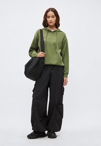 Zelená mikina s kapucňou a šnúrkami, čierne cargo nohavice s vreckami a čierne tenisky. Model drží veľkú čiernu tašku; jednoduché pozadie.