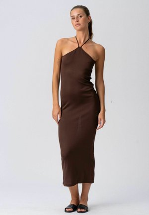 Žena stojaca, oblečená v hnedých midi šatách bez rukávov s halterneck výstrihom a čiernych šľapkách na jednoduchom svetlom pozadí.