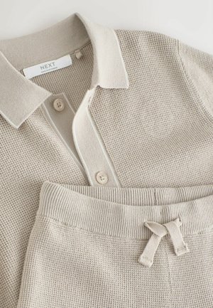 Bēša trikotāžas polo krekls ar teksturētu rakstu un divām pogām, kopā ar atbilstošām ribotām biksēm ar auklu jostasvietā.