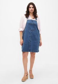 Denim overall obleka v modri barvi z nastavljivimi naramnicami, sprednjim žepom in dolžino do sredine stegen. Nosi se z bež platformnimi sandali.