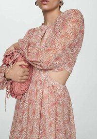 Dlhé, splývavé šaty v svetloružovej farbe s paisley vzorom, s vykrojenými bokmi a nadýchanými rukávmi. Modelka drží textúrovanú ružovú tašku.
