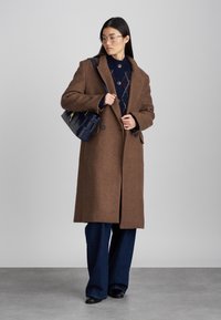 Cappotto di lana marrone con ampi risvolti, bottoni anteriori e tasche laterali, abbinato a un maglione a rombi blu e jeans denim a gamba larga.