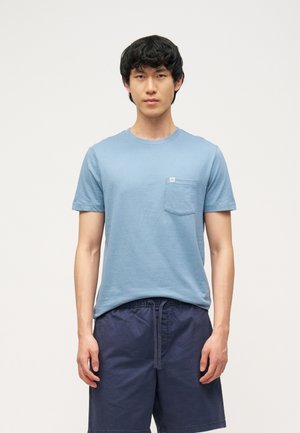 Młody mężczyzna w jasnoniebieskim, krótkim rękawie koszulce z kieszenią z przodu i ciemnoniebieskich spodenkach z trokiem, stojący na jednolitym tle.