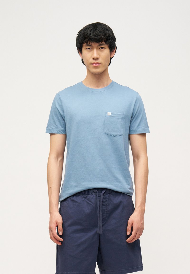 Νεαρός άνδρας που φοράει ανοιχτό μπλε κοντομάνικο μπλουζάκι με μπροστινή τσέπη και σκούρες μπλε βερμούδες με κορδόνι, στέκεται μπροστά σε ένα απλό φόντο.