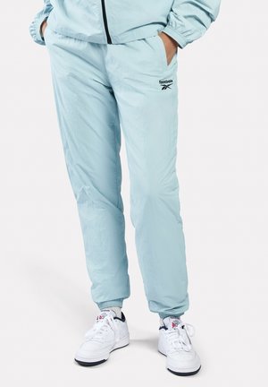 Persoon draagt lichtblauwe Reebok trainingsbroek met handen in de zakken en witte Reebok sneakers op een witte achtergrond.