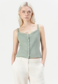 Top corto a coste verde chiaro con spalline sottili e apertura a bottoni, caratterizzato da un design aderente e una texture morbida. Indossato con pantaloni beige chiaro.