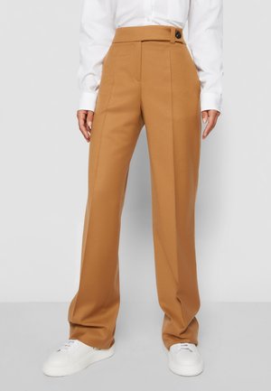High-waist wide-leg camel pants with central crease, worn with white buttoned shirt and white sneakers.