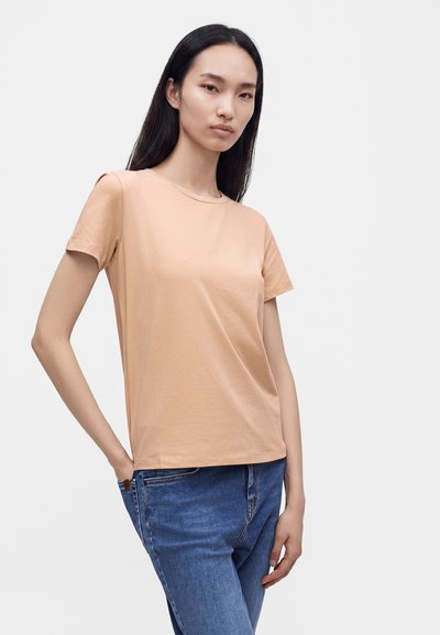 Femme aux longs cheveux noirs portant un t-shirt à manches courtes marron clair et un jean bleu, debout avec une main dans la poche devant un fond uni.