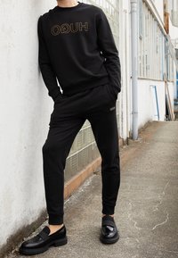 Zwarte sweatshirt met gouden "HUGO" logo, gecombineerd met zwarte joggers. De outfit heeft een strak ontwerp en wordt gecompleteerd door glanzende zwarte loafers.