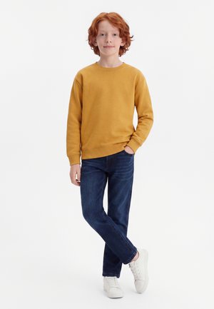 Rødhåret barn med hånd i senneps-hættetrøjes lomme, iført blå jeans og hvide sneakers, stående med det ene ben over det andet.