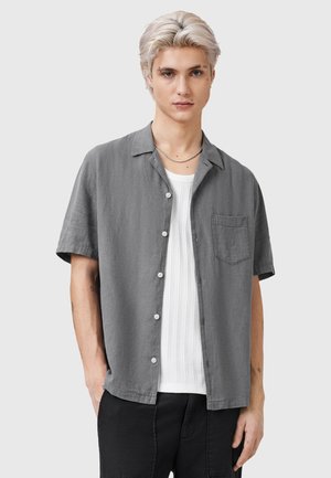 Camicia grigia a maniche corte con bottoni, dotata di taschino sul petto, realizzata in tessuto leggero, indossata sopra una maglietta bianca a trama. Design del colletto semplice.