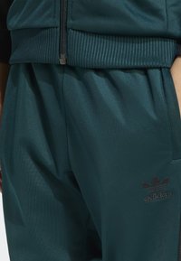 Turkosa träningsbyxor i ett slätt tyg, med ett elastiskt midjeband och en liten svart Adidas-logga på underbenet.