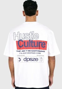 White t-shirt featuring bold red and blue text on the back, with graphic details and a relaxed fit design. Short sleeves and round neckline.
