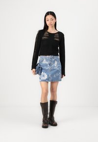 Črni pleteni pulover z uničeno teksturo, v kombinaciji z cvetličnim denim krilom. Model nosi rjavice z višino do kolen. V roki drži modro tkaninsko dodatno opremo.