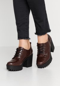 Chaussures en cuir synthétique crocodile marron avec une épaisse semelle en caoutchouc noir et un laçage sur le devant. Elles présentent un bout arrondi et un talon épais.