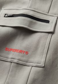 Grijze kleding met een ritssluiting zak en een rood "SUPERDRY" logo. De stof heeft een zachte textuur met zichtbare stikdetails.