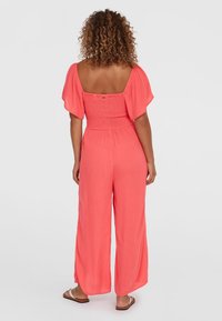 Korallfärgad byxdress med korta puffärmar, smockad livstycke, vida ben och lättviktsmaterial. Har en fyrkantig rygg.
