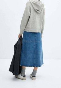 Grijze hoodie met een capuchon, gedragen over een blauwe denim maxi rok. Witte sneakers, grijze sokken en een donker gestreepte jas in de linkerhand.