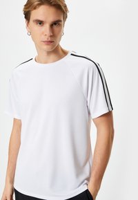 Hvid atletisk t-shirt med sorte sidelister, korte ærmer og rund halsudskæring. Fremstillet af et glat, åndbart stof.