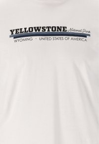 Valkoinen puuvillapaita, jossa on rohkealla mustalla tekstillä "YELLOWSTONE Kansallispuisto". Sen alla on pienemmällä fontilla tekstit "WYOMING" ja "YHDYSVALLAT".