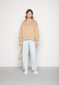 Sweatshirt com capuz castanho claro e logo branco, combinado com calças de ganga largas azul claro. Modelo segura uma bolsa bege e está de pé contra um fundo liso.