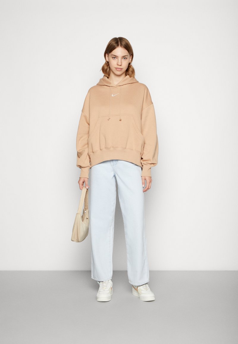 Jasnobrązowa bluza z kapturem z białym logo, w połączeniu z jasnoniebieskimi, szerokimi spodniami. Modelka trzyma beżową torbę, stoi na jednolitym tle.