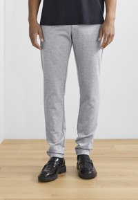 Grå joggingbukser med elastisk talje, sidelommer, tilspidsede ben og en blød tekstur, kombineret med sorte loafers på et trægulv.