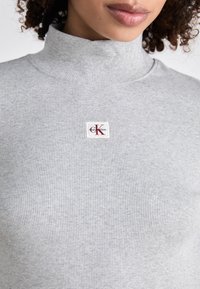 Gray ribbed turtleneck sweater made of soft fabric, featuring a small white and red Calvin Klein logo patch on the front center.
