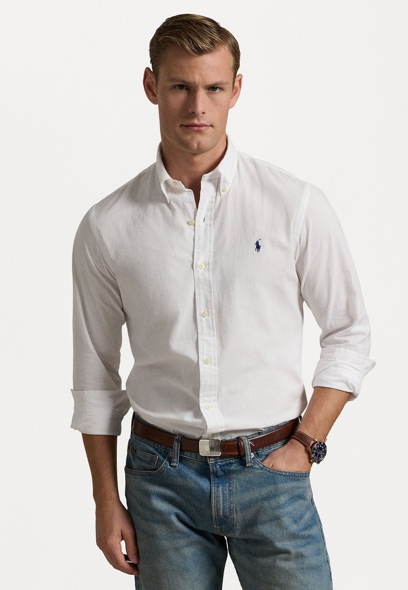 Camisa branca de botões com uma textura discreta, apresentando um logotipo azul-marinho no lado esquerdo do peito, combinada com jeans de denim azul e um cinto castanho.