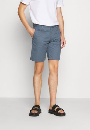 Męskie krótkie spodenki do kolan w jasnoniebieskiej bawełnie z płaskim przodem, dwoma bocznymi kieszeniami i zapięciem na guzik; zestawione z czarnymi sandałami.