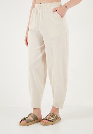 Žena nosiace voľné béžové nohavice s vysokým pásom a elastickým opaskom a olivové sandále so zlatými dvojitými prackami. Jedna ruka v vrecku.