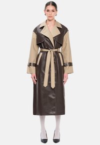 Trench-coat en cuir marron et beige avec larges revers, taille ceinturée et manches longues, présentant une longueur mi-mollet et une texture brillante.
