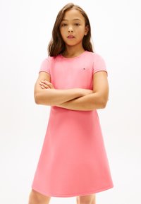 Κορίτσι με μακριά καστανά μαλλιά και σταυρωμένα τα χέρια, φορώντας ροζ φόρεμα με κοντά μανίκια, στέκεται μπροστά σε απλό λευκό φόντο.