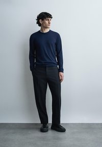 Jeune homme debout, les mains dans les poches, portant une chemise bleu marine à manches longues, un pantalon noir et des chaussures noires, regardant sur le côté.