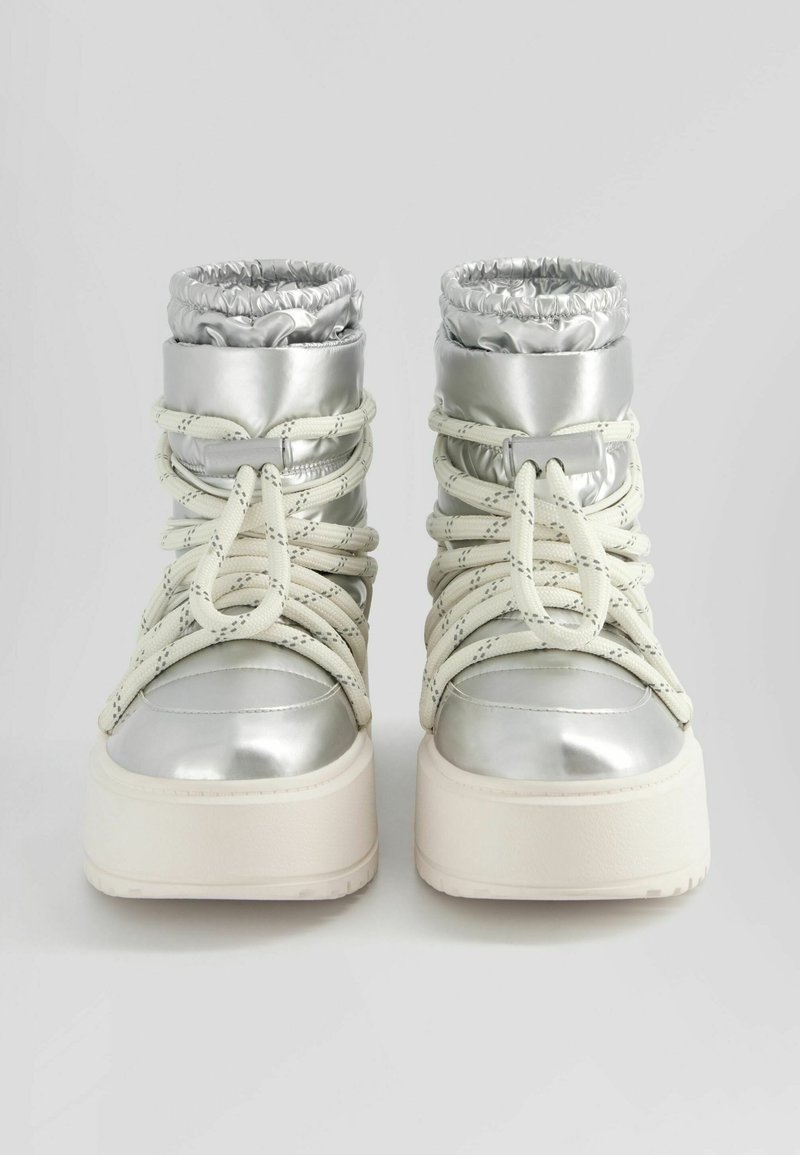 Silberne, metallische High-Top-Stiefel mit elastischen Bündchen, auffälligen Schnürsenkeln und einer dicken weißen Plateausohle. Sie haben eine glatte Textur.