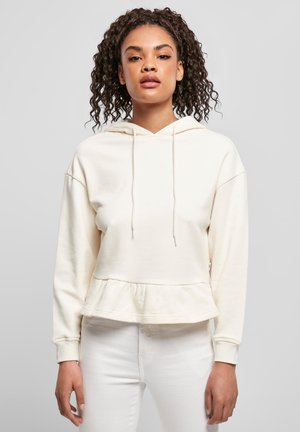 Young woman with curly hair wearing a cream hoodie with drawstrings and ruffled hem, paired with white pants, against a plain background.