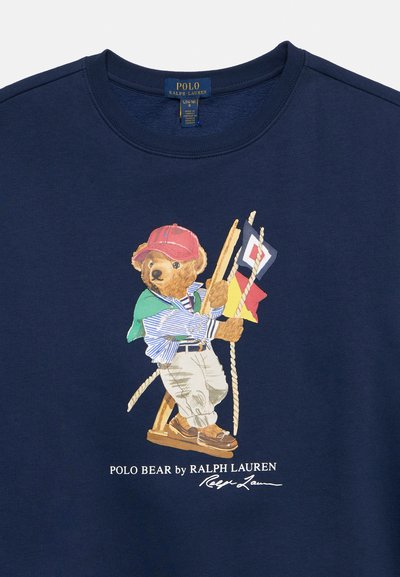 Karu punases mütsis ja triibulises särgis ronib köite peal, hoides mere lippe tumesinisel Polo Ralph Laureni särgil.