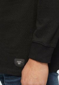 Chemise noire à manches longues avec texture côtelée, comportant une étiquette en bas à gauche affichant le nom et le logo de la marque.