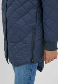 Marinblå kviltad jacka med diamantmönster, försedd med sidoglittring och stickade detaljer vid ärmarna. Materialet verkar mjukt med en lätt lyster.