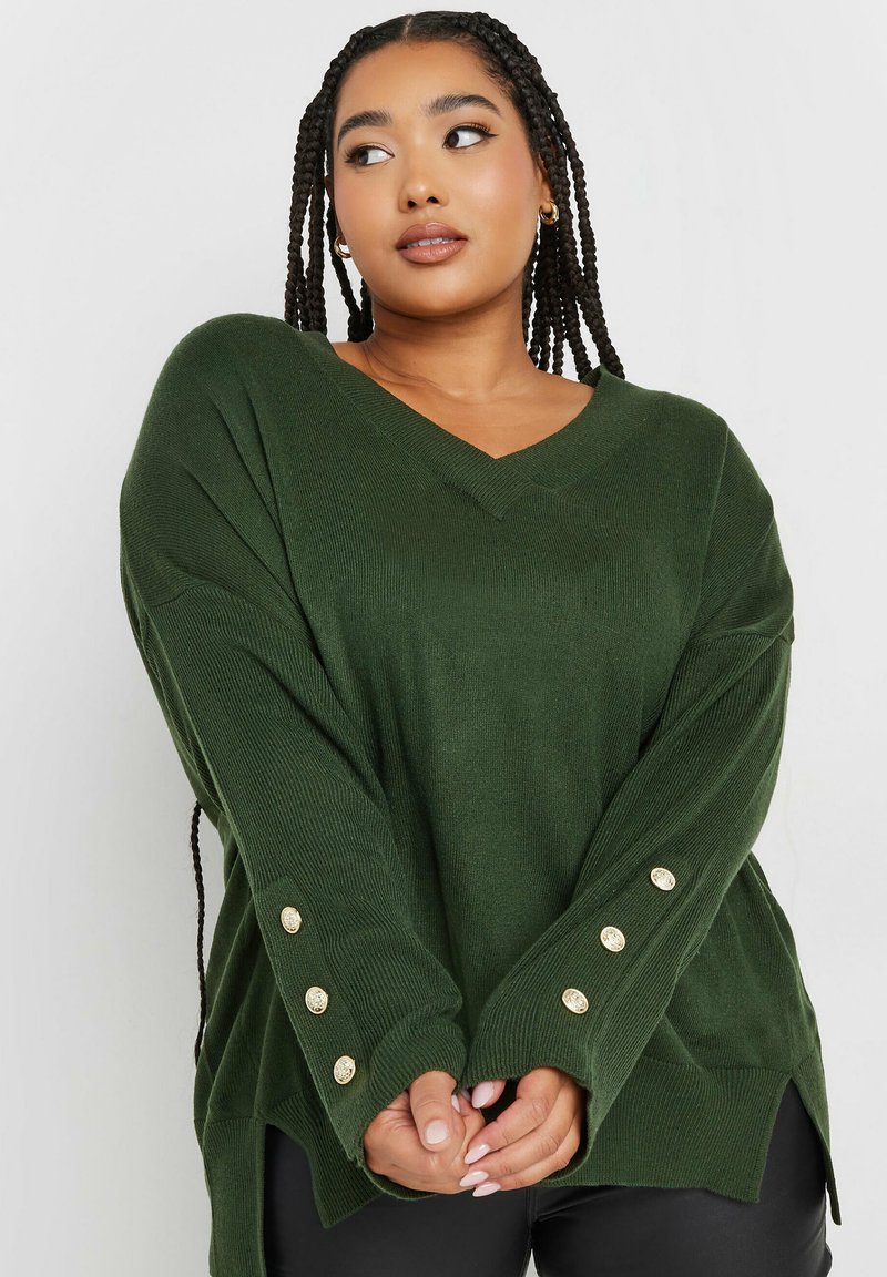 Green knit sweater with a V-neck, oversized fit, ribbed texture, and gold button accents on sleeves. Side slits add a relaxed silhouette.